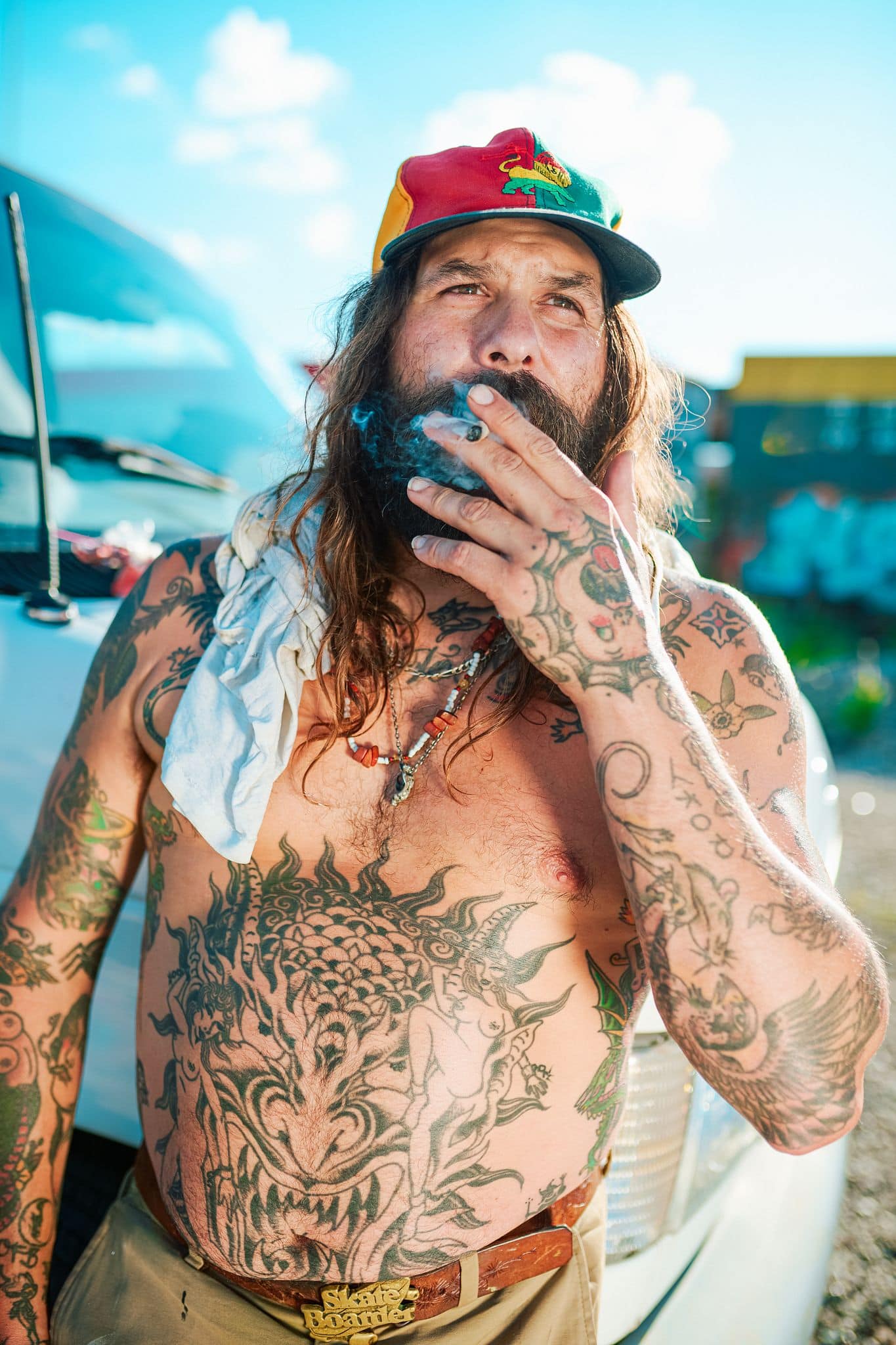Tattooed man smoking cannabis outdoors with a colorful cap.